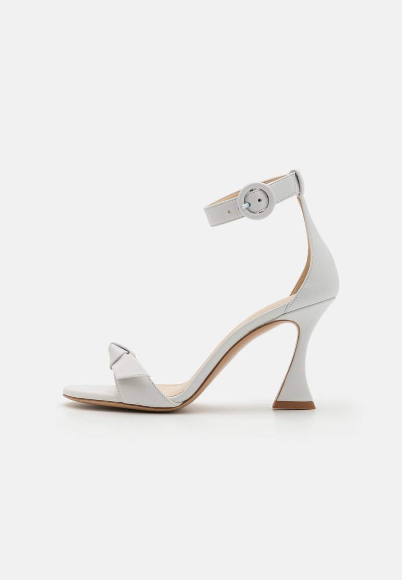 Sandalia de tacón blanca de cuero con un tacón afinado, correa en el tobillo con hebilla circular y tira frontal con un diseño retorcido. Textura suave.
