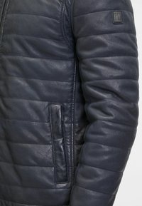 Giubbotto in pelle blu navy con design trapuntato, dotato di chiusura con zip, tasche laterali e dettagli testurizzati sulle maniche.