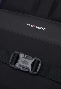 Il materiale dello zaino nero presenta una texture liscia con un'area imbottita. Evidente branding FLEXVENT e una fibbia regolabile grigia.