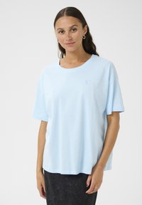 Light blue cotton t-shirt with short sleeves, round neckline, and subtle heart embroidery on the chest, paired with a dark skirt.
