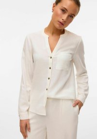 Blusa bianca a maniche lunghe con una vestibilità rilassata, chiusura frontale con bottoni e una tasca sul petto. Tessuto morbido e lucentezza sottile. Bottoni color oro.