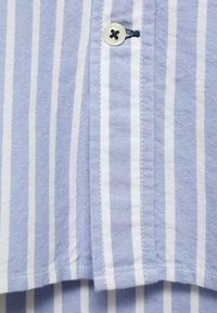 Tessuto a righe azzurro chiaro e bianco con un bottone e dettagli di cucitura. La texture appare tessuta, evidenziando l'abbigliamento casual.