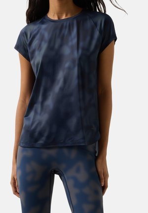 Kvinde iført en mørkeblå, kortærmet sportsbluse og matchende mønstrede leggings, stående mod en ensartet lys baggrund.