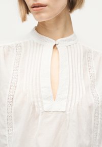 Femme portant un chemisier blanc avec des plis verticaux, une bordure en dentelle aux manches et un col en goutte d'eau boutonné.