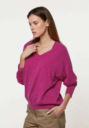 Femme aux cheveux longs bruns portant un pull ample à col en V magenta et un pantalon beige, une main touchant le col et l'autre dans la poche.
