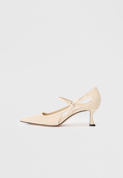 Talons en cuir beige avec bout pointu, détails ajourés sur les côtés et petit nœud décoratif. Dotés d'un talon fin et bas ainsi que d'une texture lisse.