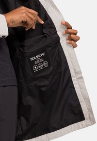 Lichtgekleurde waterdichte jas met een interieur dat een zwarte zak bevat met een TEXAPORE-label, dat de waterdichte en ademende eigenschappen benadrukt.