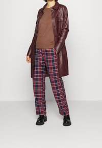 Pull en tricot marron, manteau en cuir bordeaux et pantalon à carreaux avec des lignes rouges, bleues et blanches. Des chaussures à plateforme noires complètent la tenue.