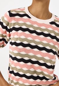 Zbliżenie na osobę noszącą sweter z kolorowego dzianinowego materiału w faliste paski w odcieniach różowego, czarnego, oliwkowego i kremowego, w połączeniu z cienką złotą naszyjnikiem.
