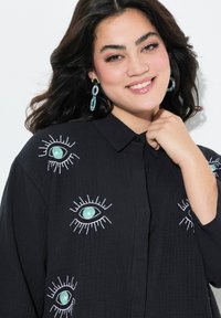 Zwarte blouse met knopen, versierd met grote geborduurde oogontwerpen in teal en wit, met een gestructureerde stof. Het model heeft lang, donker haar en draagt oorbellen.