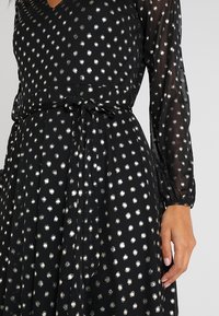Schwarzes Kleid mit silbernen Punkten, langen durchsichtigen Ärmeln, einem V-Ausschnitt und einer gebundenen Taille. Der Stoff hat eine weiche, fließende Textur.