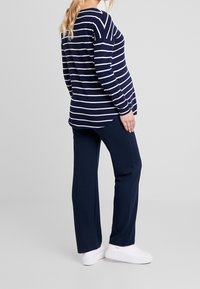 Femme enceinte debout de profil portant un haut à manches longues rayé bleu marine et blanc, un pantalon bleu marine, et des baskets blanches sur un fond uni.