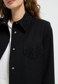 Black jacket featuring a textured beaded collar and pocket detail, with gold snap buttons and a smooth fabric finish.