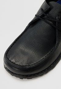 Chaussure en cuir noir avec une surface texturée, bout rond, détails cousus et lacets en haut. La semelle est en caoutchouc pour une adhérence et une durabilité accrues.