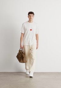 Hvid t-shirt med korte ærmer og et rødt oval design, parret med beige bukser og hvide sneakers. Bærer en brun tekstureret taske.