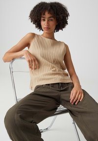 Young woman with curly hair seated on a clear chair, wearing a beige ribbed sleeveless top and loose dark olive pants.