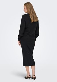 Ženska z ravnimi dolgimi lasmi nosi črni pleteni pulover, rebrasto midi krilo in črne sandale z visokimi petami, stoji obrnjena levo.