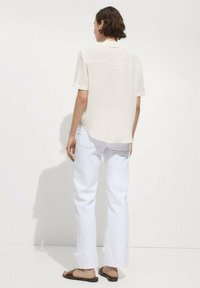 Cream short-sleeve blouse with a relaxed fit, paired with white straight-leg jeans and brown slide sandals, seen from behind.