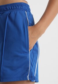 Modré sportovní šortky z lehkého materiálu, s elastickým páskem, bočními kapsami a bílými akcentovými pruhy podél švů.