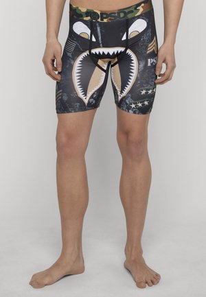 Mann trägt schwarze und tarnfarbene Kompressionsshorts mit Haigesicht und militärischen Aufnähern, steht barfuß vor einem weißen Hintergrund.