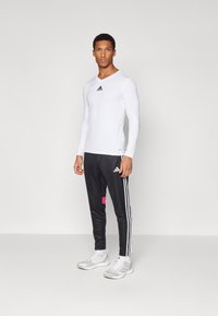 Langärmliges, weißes Sporthemd mit schwarzem Adidas-Logo, kombiniert mit schwarzen Hosen, die drei weiße Streifen und einen rosa Akzent aufweisen. Sportschuhe.