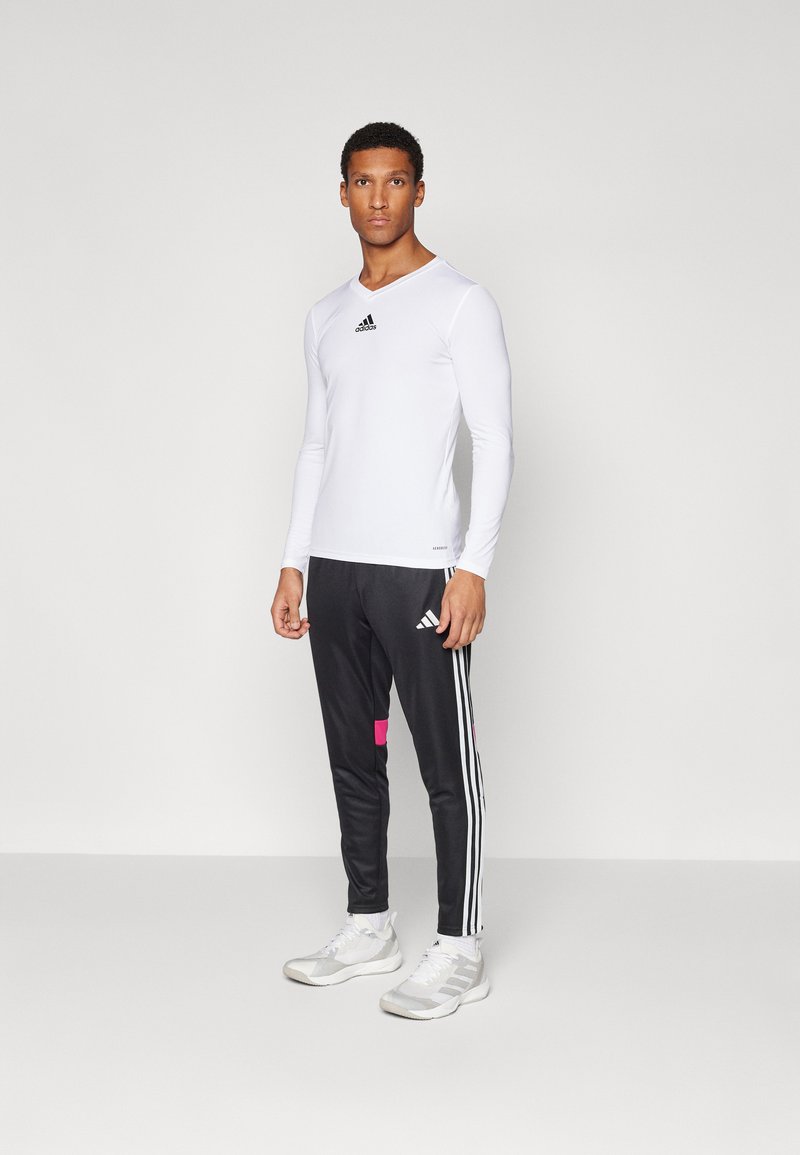 T-shirt de sport à manches longues blanc avec un logo Adidas noir, associé à un pantalon noir avec trois bandes blanches et un accent rose. Chaussures de sport.