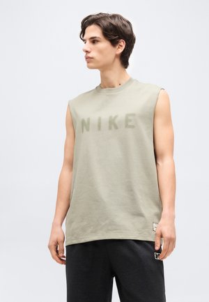 Νεαρός άνδρας με προσανατολισμό προς τα αριστερά, φορώντας μπεζ αμάνικη μπλούζα Nike και σκούρο παντελόνι, σε απλό ανοιχτό φόντο.