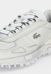 Baskets blanches avec une semelle texturée, un logo crocodile métallique sur le côté et des lacets blancs passés dans des œillets métalliques.