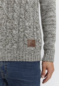 Gebreide trui met een kabelpatroon in grijs en bruintinten, geribde manchetten en een bruine labelpatch aan de voorkant. Draagt met donkere jeans.