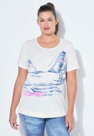 Kobieta z związanymi włosami, ubrana w białą koszulkę z nadrukiem niebieskich gór i różowej ryby, w połączeniu z niebieskimi dżinsami, stoi na białym tle.