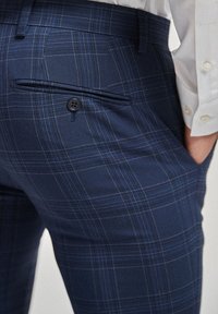 Gros plan sur un pantalon à carreaux bleu marine avec une poche arrière boutonnée et une manche de chemise blanche avec des boutons.