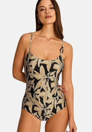 Femme portant un maillot de bain une pièce noir avec un motif de feuilles tropicales beige et des bretelles fines, debout devant un fond uni.