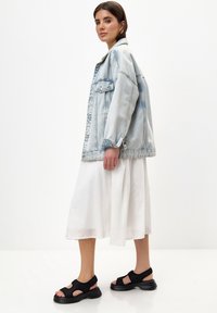 Světle modrá oversized džínová bunda, bílá splývavá šaty a černé masivní sandály s výřezy. Jednoduchý, moderní design s čistými liniemi.