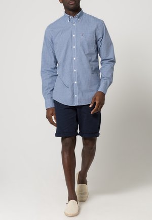 Camisa de cuadros azul claro y blanca, de botones, de mangas largas, combinada con pantalones cortos azul marino. Zapatos de lona beige sin cordones completan el atuendo.