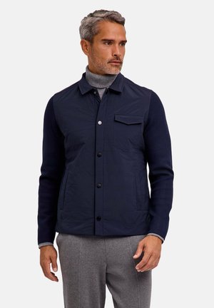 Mann mit grauen Haaren trägt eine marineblaue, geknöpfte Jacke über einem grauen Rollkragenpullover und grauen Hosen, steht und schaut zur Seite vor weißem Hintergrund.
