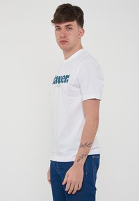 T-shirt bianco in cotone con una stampa grafica teal che riporta le scritte "lauer" e "USA" sul davanti, con maniche corte e colletto a girocollo.