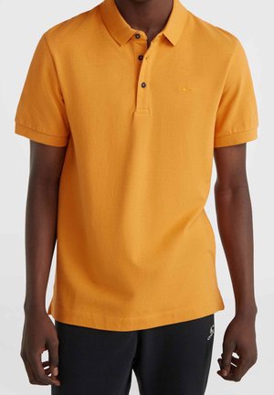 Mand iført en sennepsgul kortærmet polo-shirt med tre sorte knapper og sorte shorts mod en ensfarvet baggrund.
