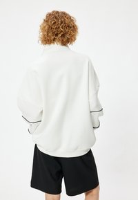 Witte oversized sweatshirt met een hoge kraag, geribde manchetten en zwarte accenten, gecombineerd met zwarte shorts, weergegeven vanuit de achterkant.