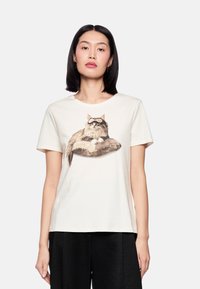 Weißes Baumwoll-T-Shirt mit einem gedruckten Bild einer langhaarigen Katze auf einem gemusterten Kissen. Kurze Ärmel und ein runder Halsausschnitt.