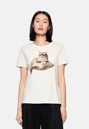 Weißes Baumwoll-T-Shirt mit einem gedruckten Bild einer langhaarigen Katze auf einem gemusterten Kissen. Kurze Ärmel und ein runder Halsausschnitt.