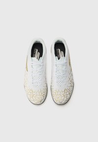Baskets de sport blanches avec un motif craquelé texturé doré. Comprend un orteil perforé, des lacets plats et un col rembourré. Semelles intérieures noires visibles.