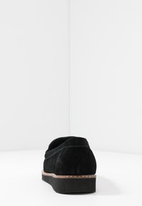 Svart mockaskor med rund tå, låg klack och kontrasterande söm längs sulan. Slät textur, minimalistisk design.