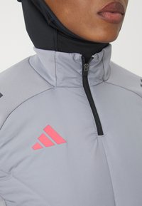 Primer plano de una persona que lleva una chaqueta gris con cremallera y un logo rojo de Adidas, con una capucha negra debajo, mostrando los labios y la barbilla.