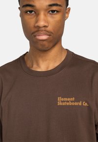 Camiseta de algodón marrón con cuello redondo, que presenta el logo "Element Skateboard Co." en naranja en el lado izquierdo del pecho. Textura suave, ajuste casual.