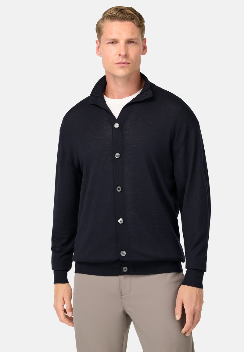 Marinfärgad cardigan, stickad textur, V-ringad krage, sex svarta knappar, ribbade ärmslut och fåll, bärs över en vit skjorta och beiga byxor.