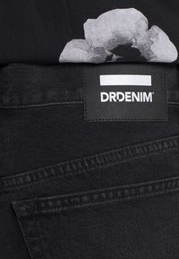Schwarzer Denim-Stoff mit sichtbaren Nähten. Enthält ein Etikett mit der Aufschrift "DRDENIM" in Weiß auf einem rechteckigen schwarzen Patch.