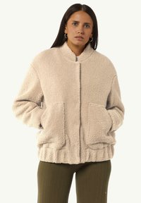Beżowa, teksturowana polarowa kurtka z wysokim kołnierzem, przednimi kieszeniami i elastycznym dołem. Krój jest lekko oversize i casualowy.