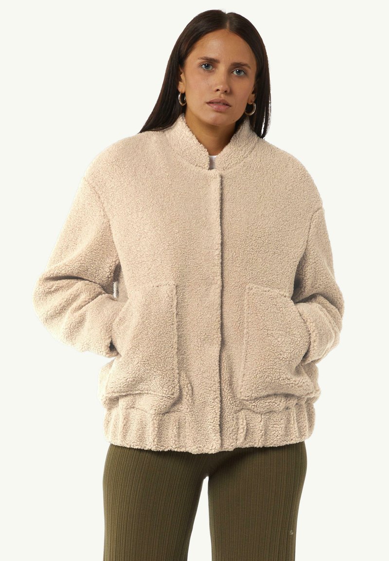 Beżowa, teksturowana polarowa kurtka z wysokim kołnierzem, przednimi kieszeniami i elastycznym dołem. Krój jest lekko oversize i casualowy.