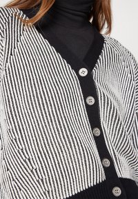 Femme portant un cardigan rayé noir et blanc boutonné sur un pull à col roulé noir, gros plan sur le haut du torse.