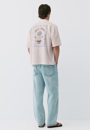 Homme portant une chemise rose clair à manches courtes avec un motif d'ananas et un texte au dos, associée à un jean bleu clair et des sandales noires.